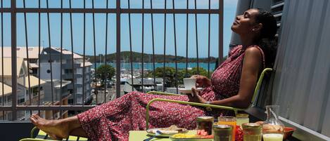 Un balcón con una mesa dispuesta para el desayuno, una persona sentada y vistas a edificios y al agua.