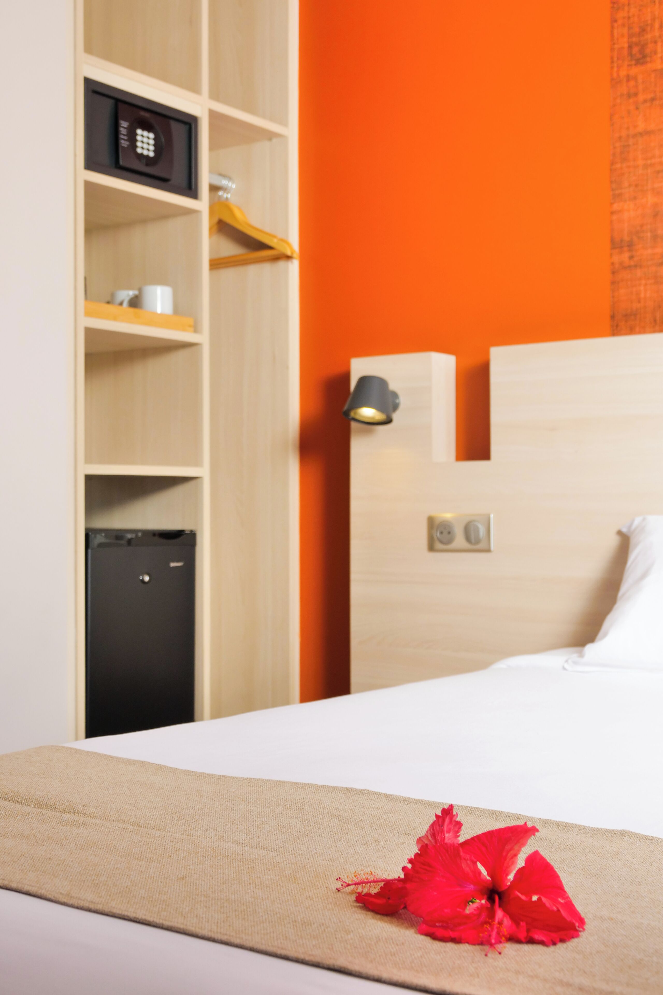 A hotel room with a bed, a minibar, a wall-mounted lamp, and a power outlet.