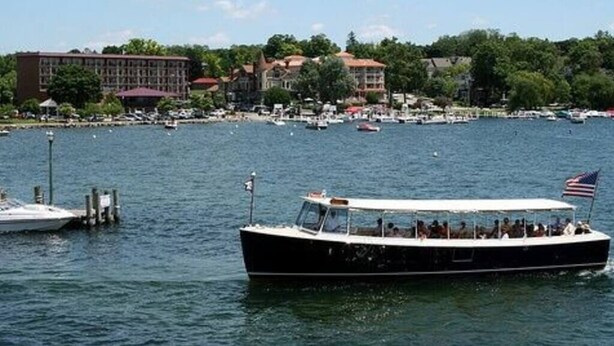 Un barco con pasajeros y una bandera, amarrado cerca de un puerto deportivo con edificios y árboles al fondo.