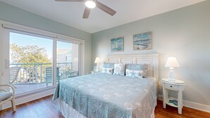 A bedroom with a large bed, two bedside tables with lamps, a ceiling fan, and a balcony with chairs.
