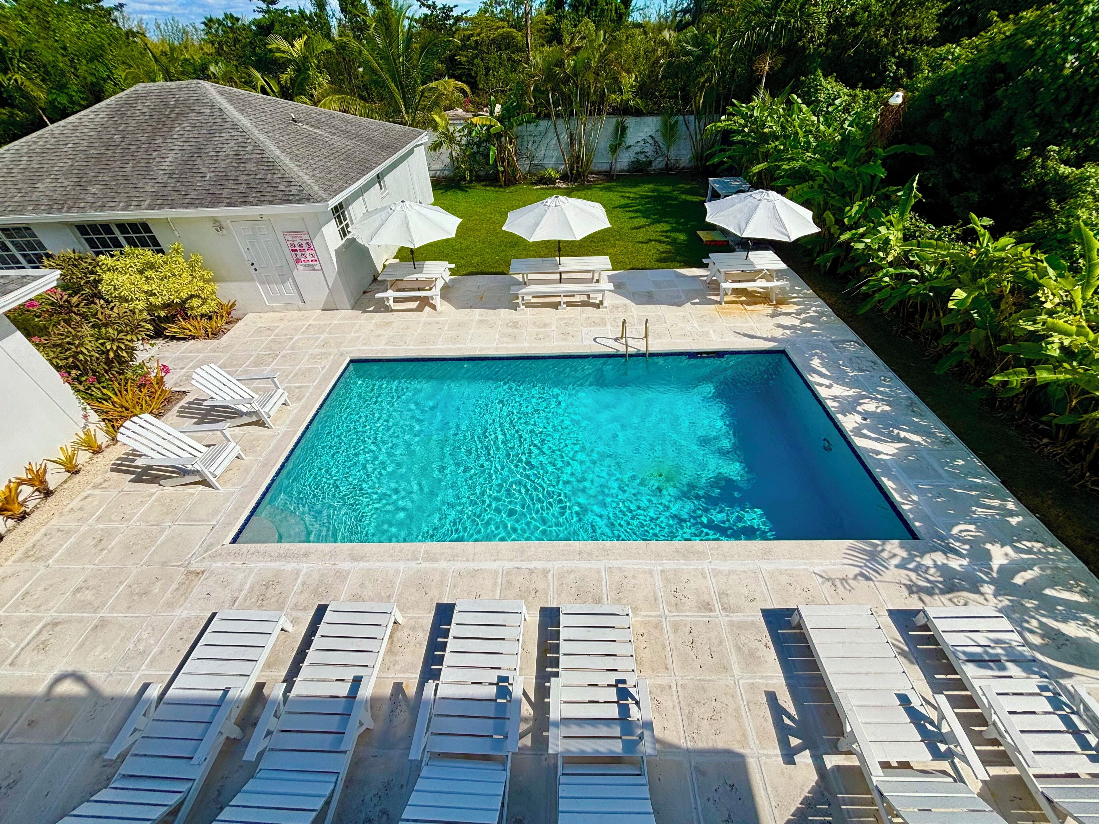 A rectangular swimming pool with clear blue water, surrounded by white lounge chairs and umbrellas, set against a lush green garden with a white building in the background.