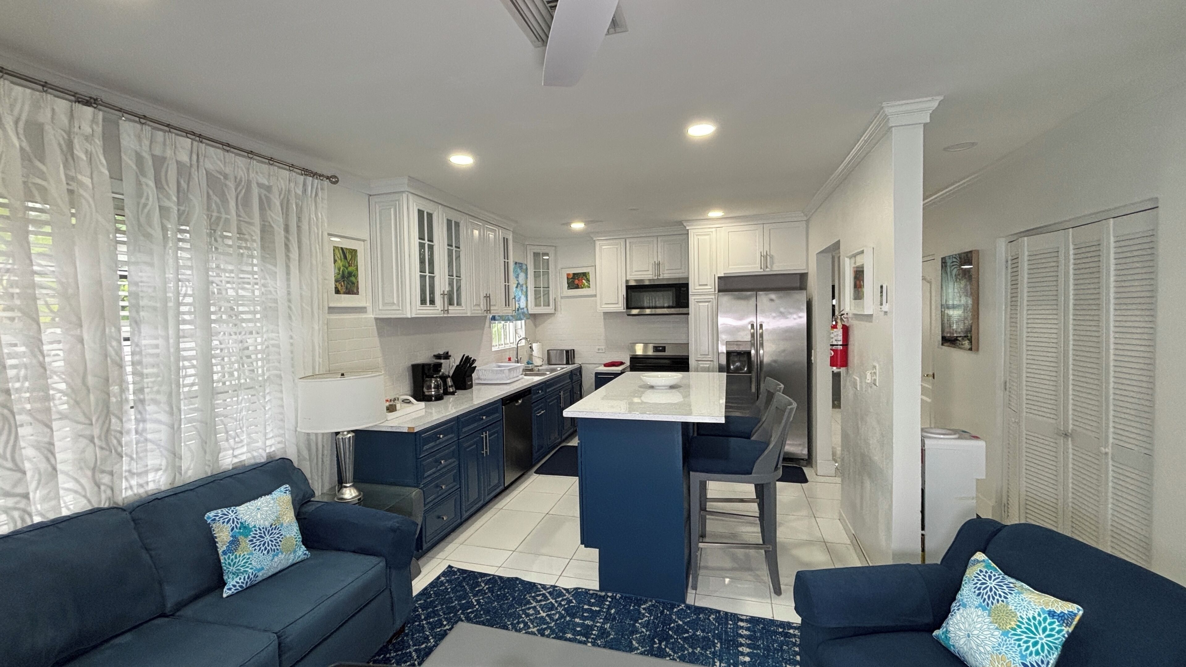 Una cocina moderna con armarios blancos, una isla central y electrodomésticos de acero inoxidable. Un salón con dos sofás azul oscuro y cojines estampados, una ventana grande y cortinas blancas.