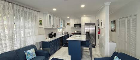 Una cocina moderna con armarios blancos, una isla central y electrodomésticos de acero inoxidable. Un salón con dos sofás azul oscuro y cojines estampados, una ventana grande y cortinas blancas.
