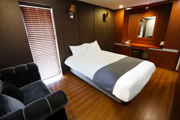 Une chambre d’hôtel avec un grand lit, un coin salon, une coiffeuse avec un miroir et une fenêtre avec des stores.