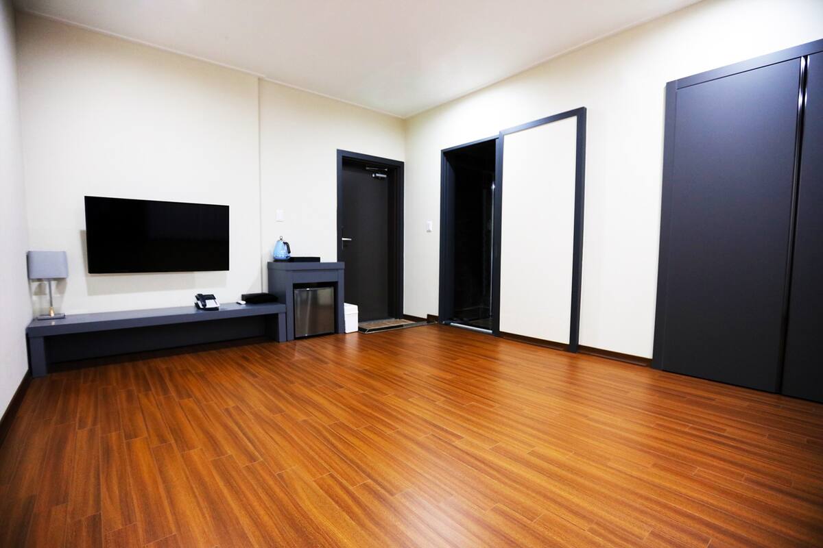 A room with a wooden floor, a flat-screen TV mounted on the wall, a small desk with a lamp, and a door leading to another room.