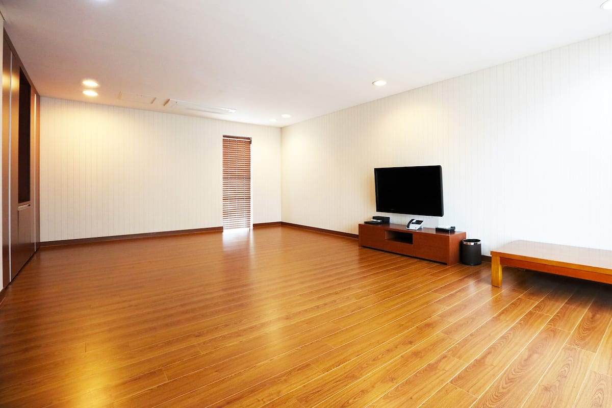 A spacious room with wooden flooring, a flat-screen TV on a wooden stand, and a sliding door with blinds.