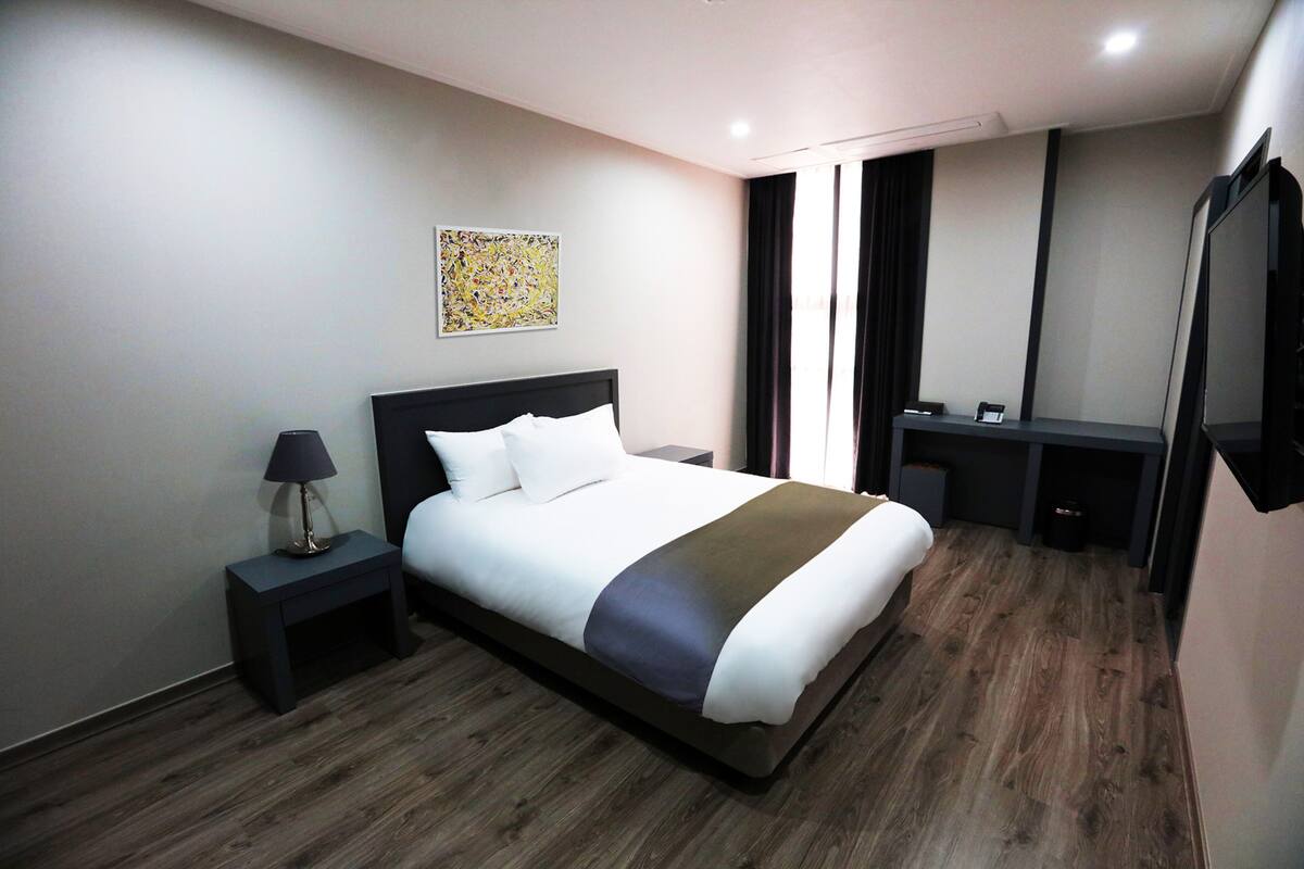 A modern hotel room with a large bed, a bedside table with a lamp, a desk with a chair, and a large mirror.