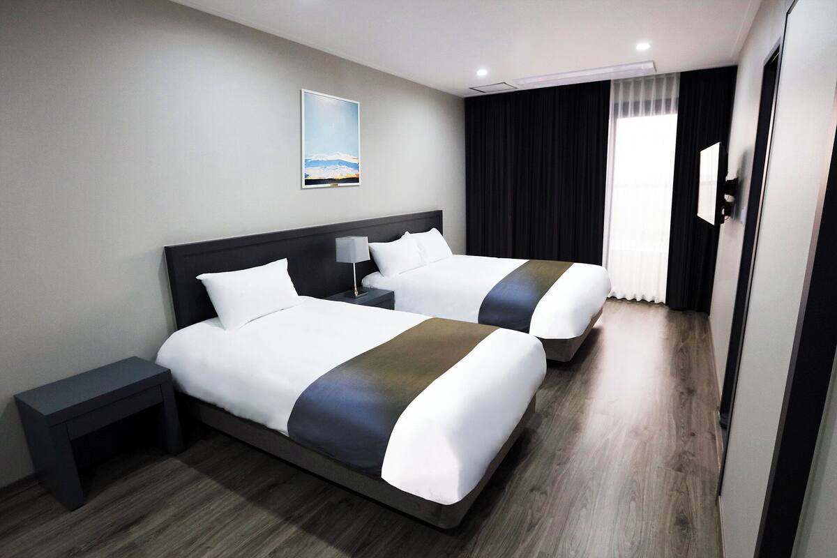 A hotel room with two beds, a nightstand, a lamp, a painting on the wall, and a window with curtains.
