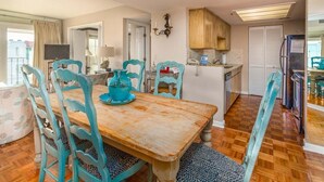 A dining area with a wooden table and turquoise chairs, a kitchen with a refrigerator and cabinets, and a television in the background. - Oceanview unit at The Beach Club.  Pool, Great Bea (Saint Simons Island)