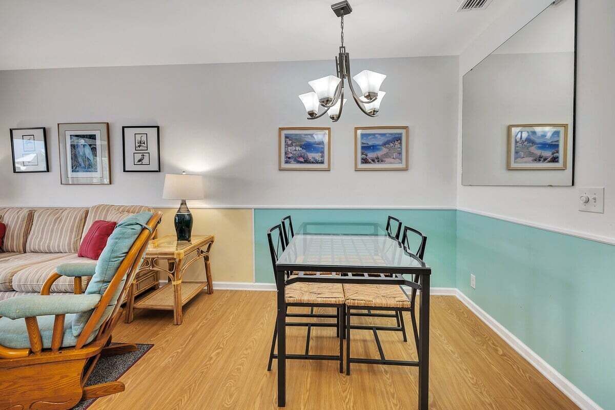A dining area with a glass-top table and chairs, a wooden chair, a lamp, and framed pictures on the walls.