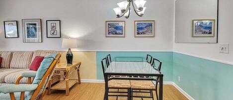 A dining area with a glass-top table and chairs, a wooden chair, a lamp, and framed pictures on the walls.
