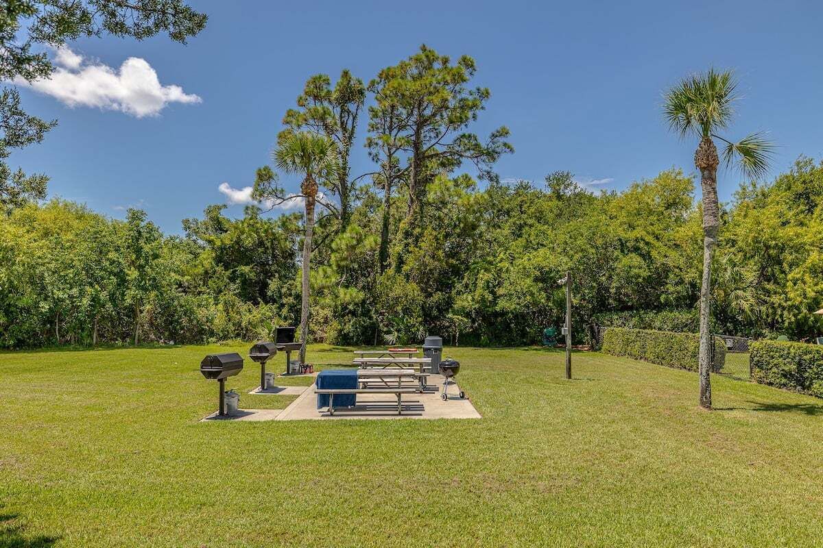 Un espace extérieur aménagé avec des tables de pique-nique, des barbecues et une pelouse verdoyante entourée d’arbres.