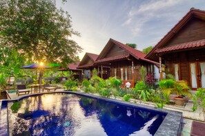 A pool area with wooden cabins, surrounded by lush greenery and a clear sky. - Asoka Bungalows (Penida Island)