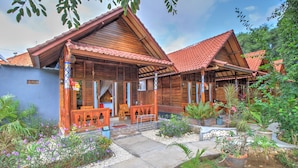 A wooden house with a tiled roof, a porch with wooden furniture, and a garden with various plants. - Asoka Bungalows (Penida Island)