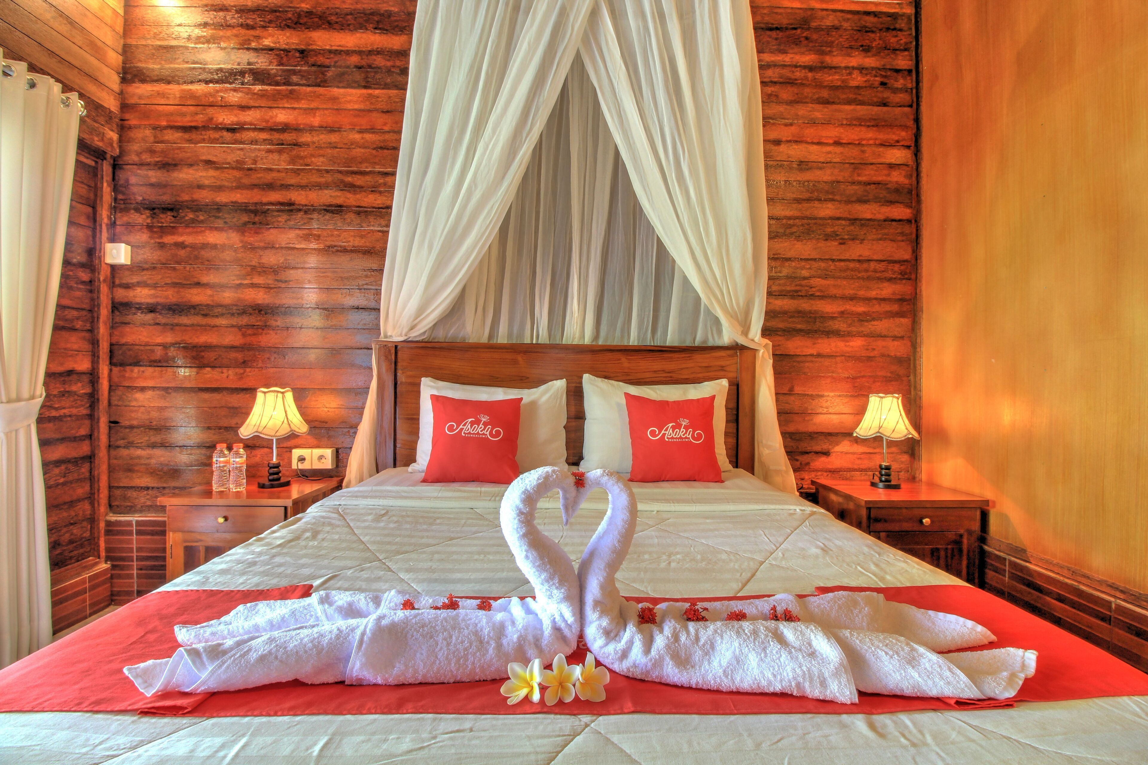 A hotel room with a wooden wall, a bed with red and white bedding, two pillows with 'Asoka' branding, a nightstand with a lamp, and a decorative towel arrangement on the bed.