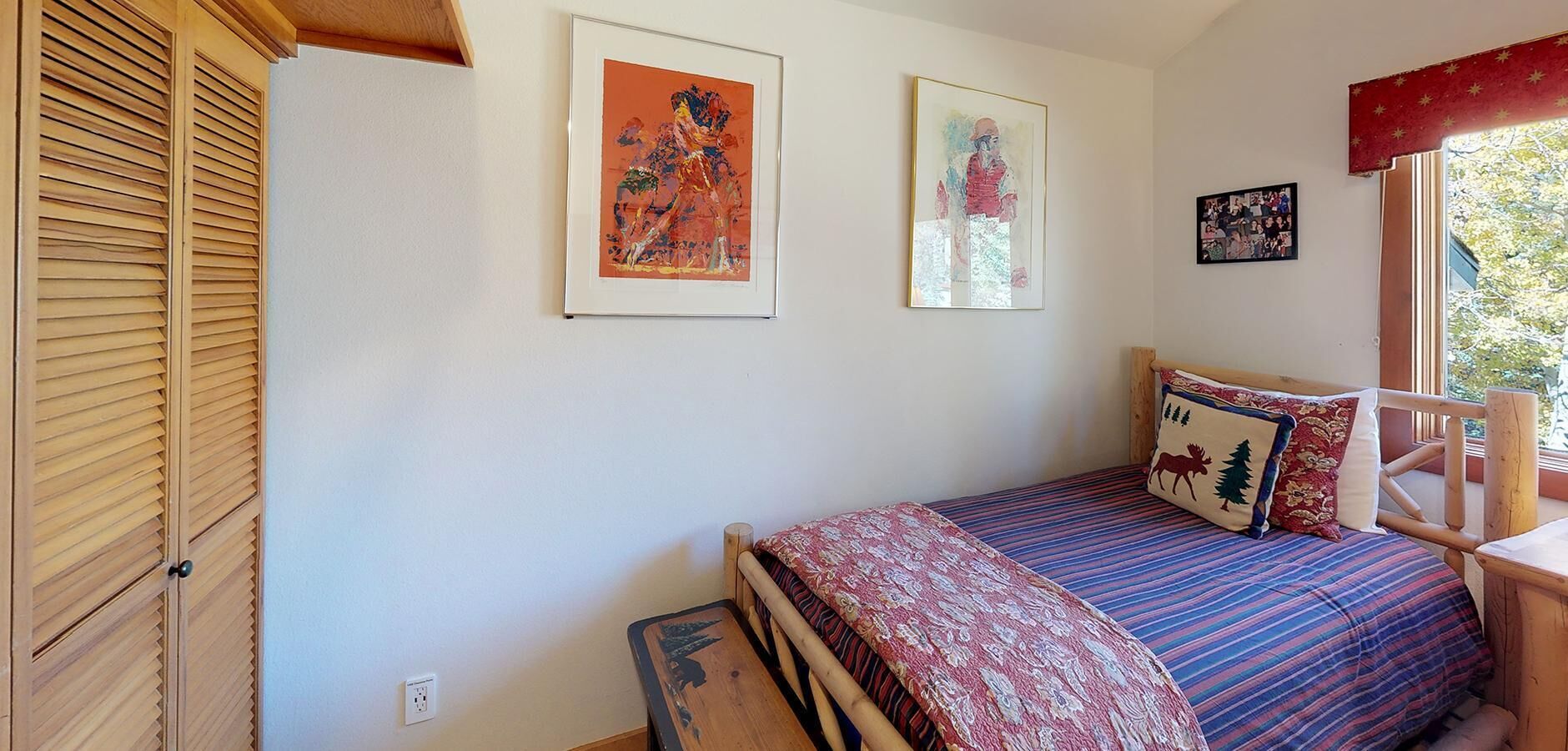 A bedroom with a bed, two framed artworks, a window with a view, and a wooden wardrobe.