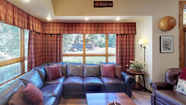 A cozy living room with a leather sectional sofa, a small side table with a lamp, and a sign reading "The Barge Inn" above a window with plaid curtains. - Elegant Creekside Condo with Scenic Views & Pool with Access to Grand Hyatt (Vail)