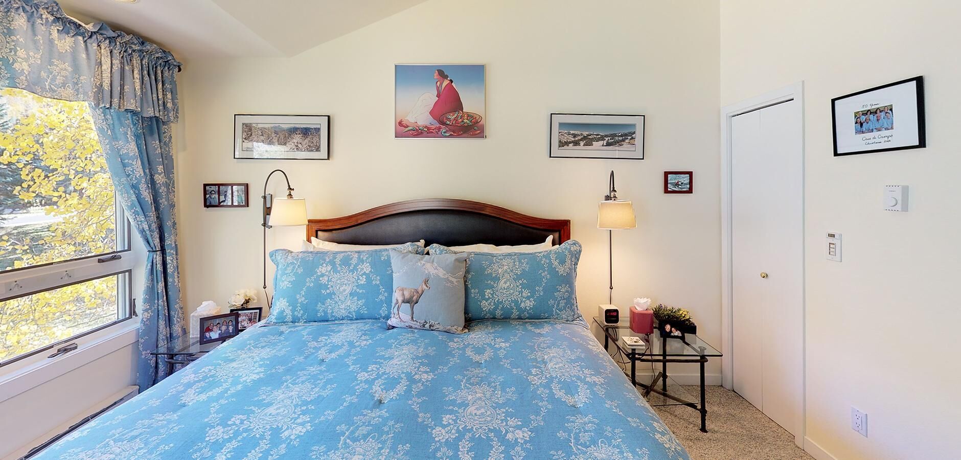 Une chambre à coucher avec un grand lit, deux tables de chevet, une commode, une fenêtre avec des rideaux et des œuvres d’art accrochées au mur.