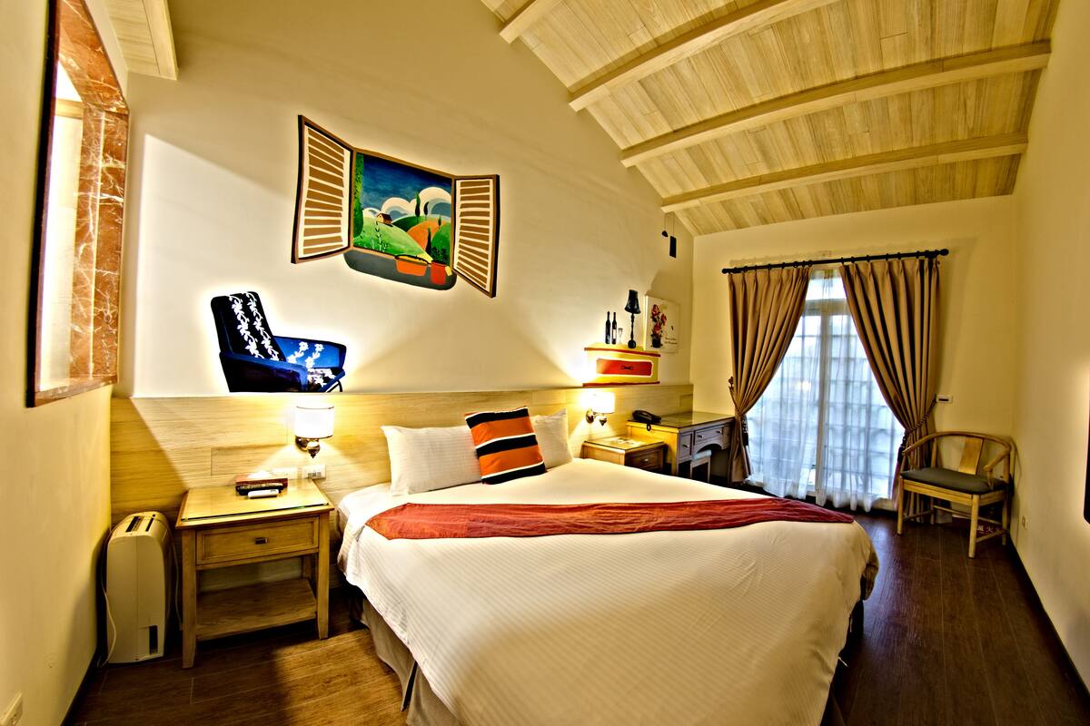 A hotel room with a large bed, a nightstand with a lamp, a chair, and a painting on the wall.