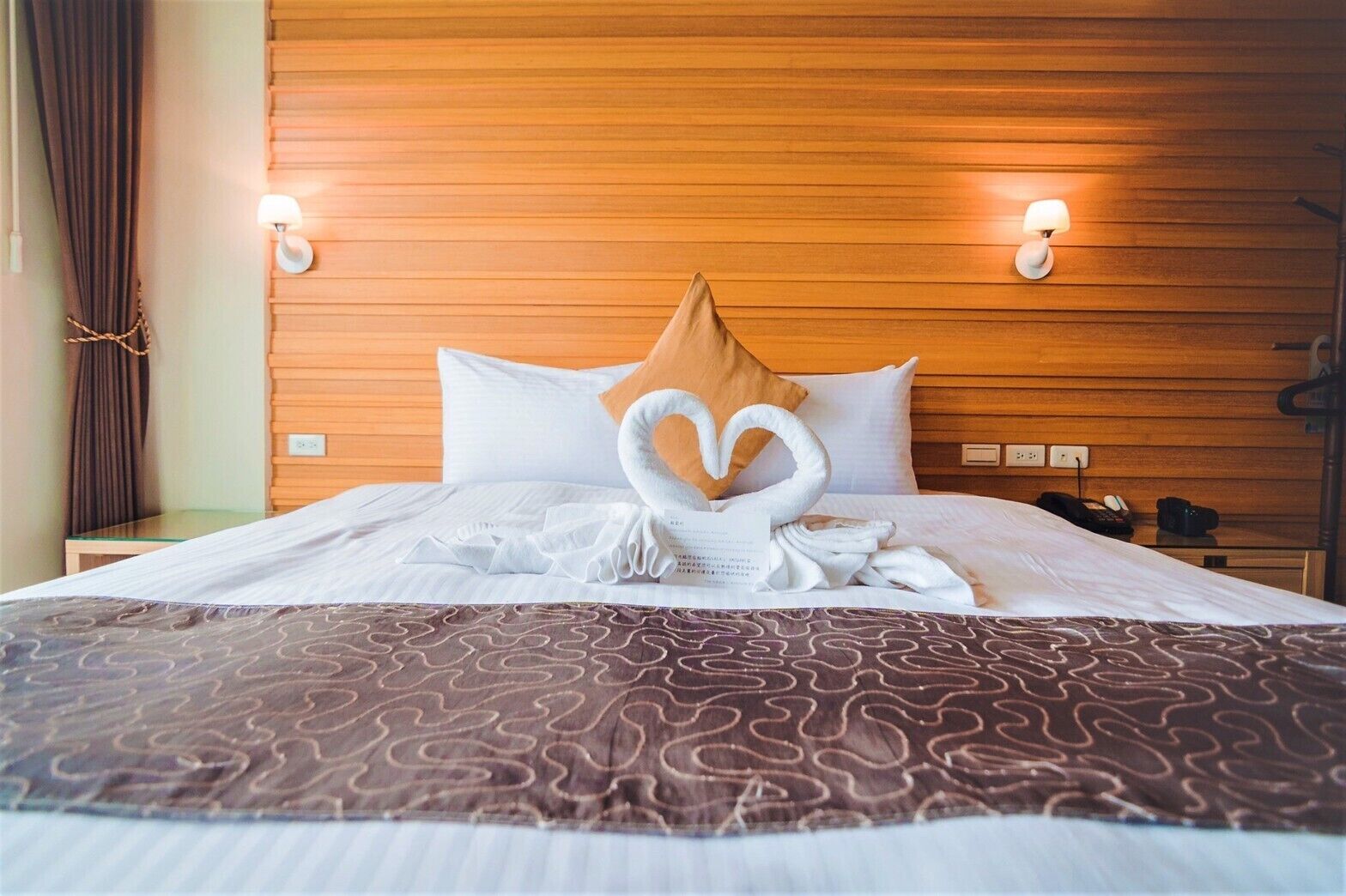 A hotel room with a wooden headboard, two bedside lamps, a bed with a heart-shaped towel arrangement, and a patterned throw blanket.