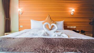 A hotel room with a wooden headboard, two bedside lamps, a bed with a heart-shaped towel arrangement, and a patterned throw blanket.