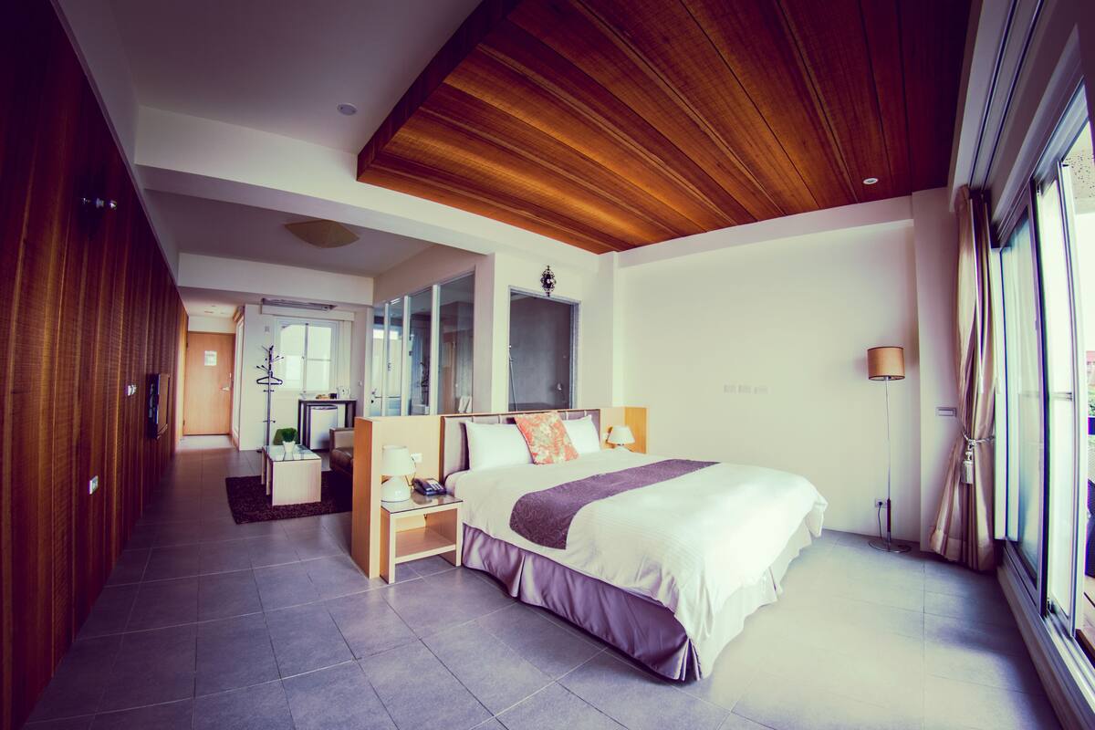 A modern hotel room with a large bed, wooden ceiling, and a view of the outdoors through a large window.