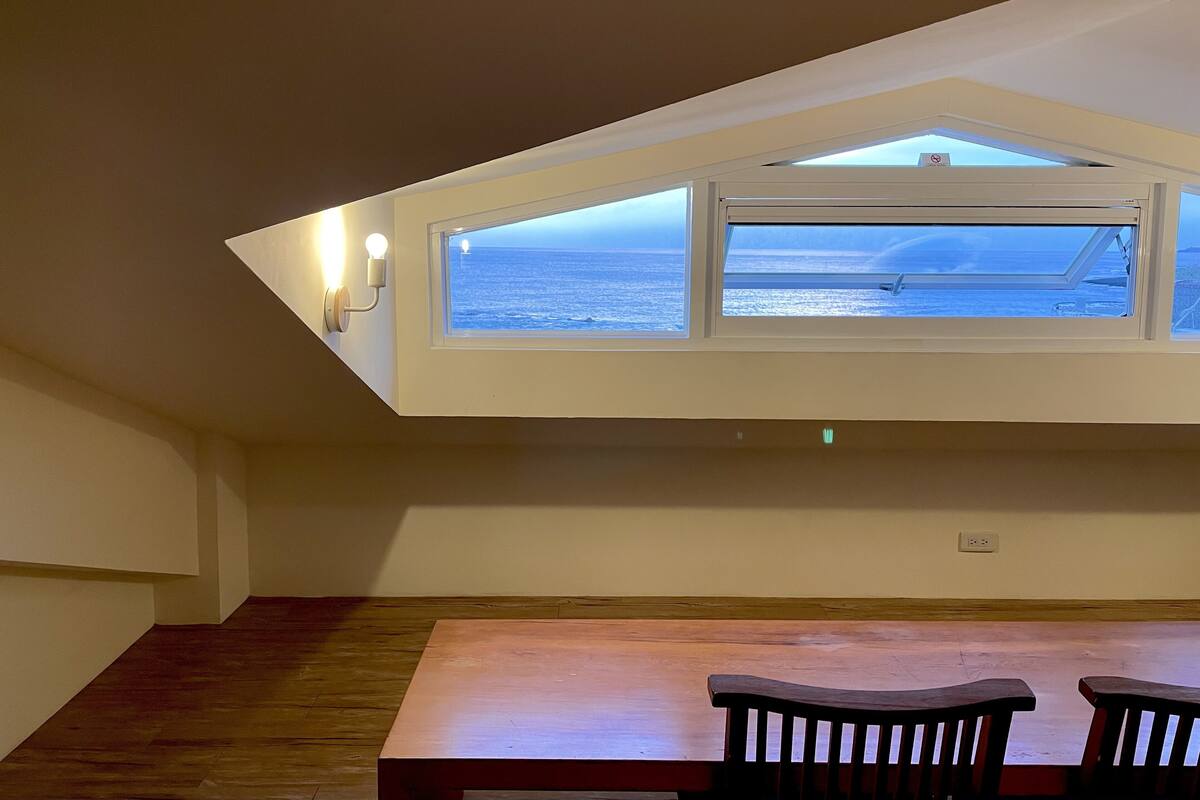 A room with a sloped ceiling, a table with two chairs, and a window overlooking the sea.