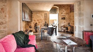 Un intérieur rustique avec des murs en pierre, un canapé rose, une table ronde recouverte de papiers et un coin repas avec des chaises et une table.