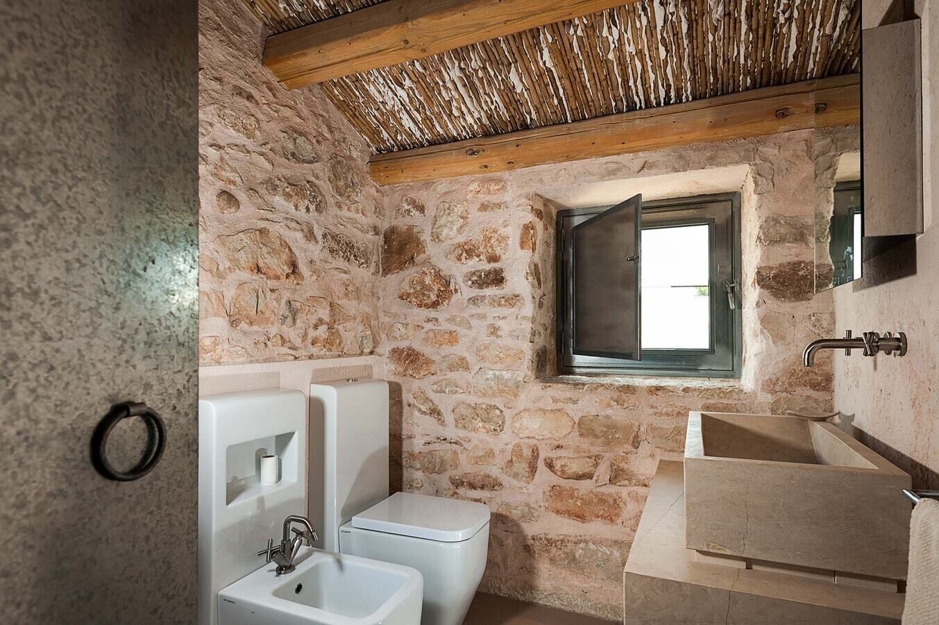 A rustic bathroom with stone walls, a wooden ceiling, a window, a toilet, and a sink.