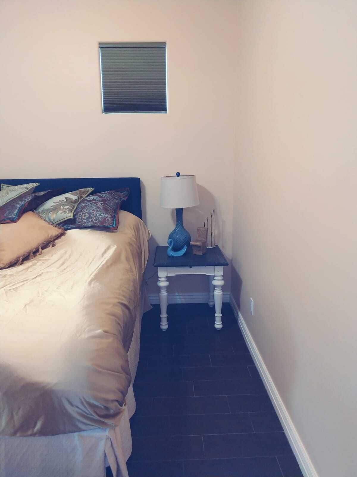 A bedroom with a bed, a bedside table with a lamp, and a wall-mounted air vent.