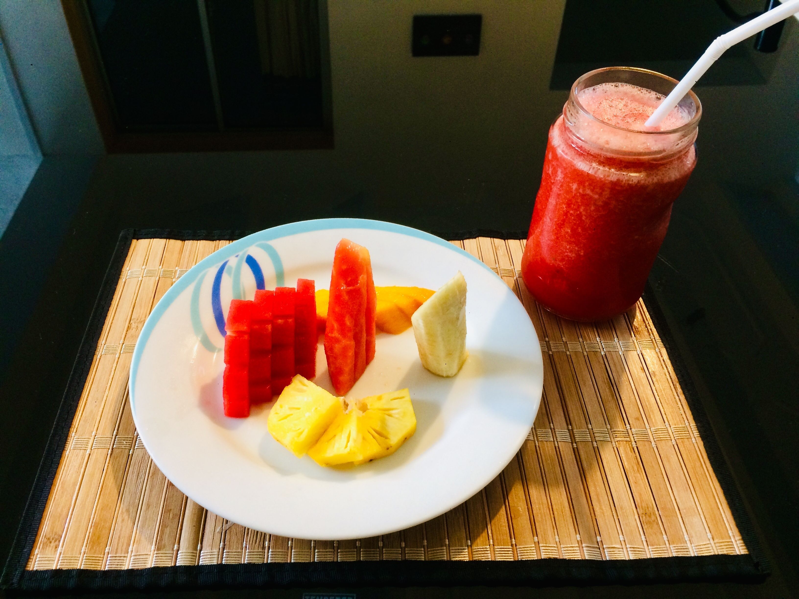 Un plato con rebanadas de sandía, piña y plátano, y un vaso de batido rojo sobre una estera de bambú.