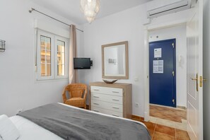 Ein Schlafzimmer mit einem Bett, einem Fernseher, einem Korbstuhl, einer Kommode, einem Spiegel, einer blauen Tür und einem Fenster mit Vorhängen.