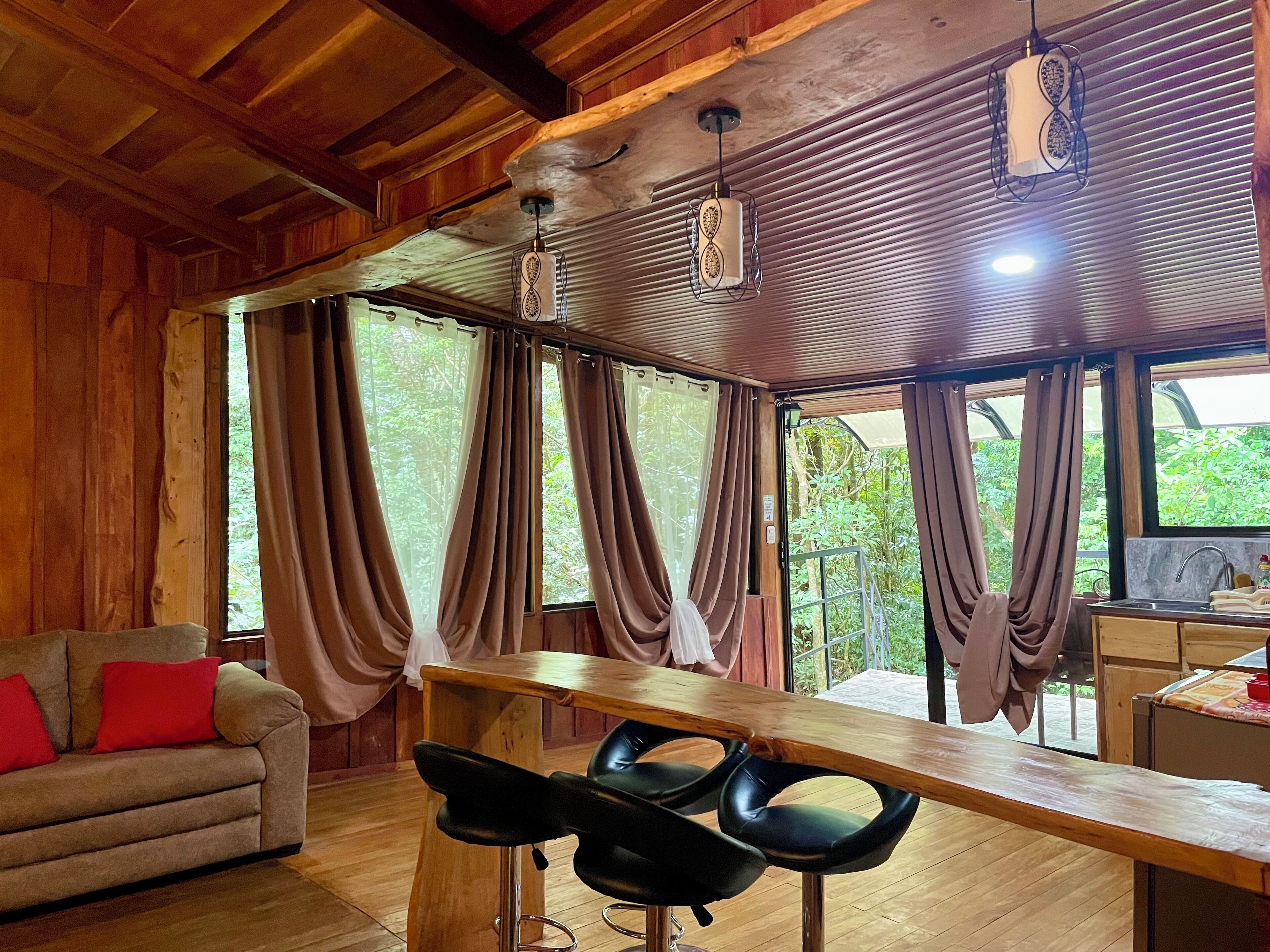 A wooden interior with a sofa, two bar stools, a wooden table, and large windows with curtains.