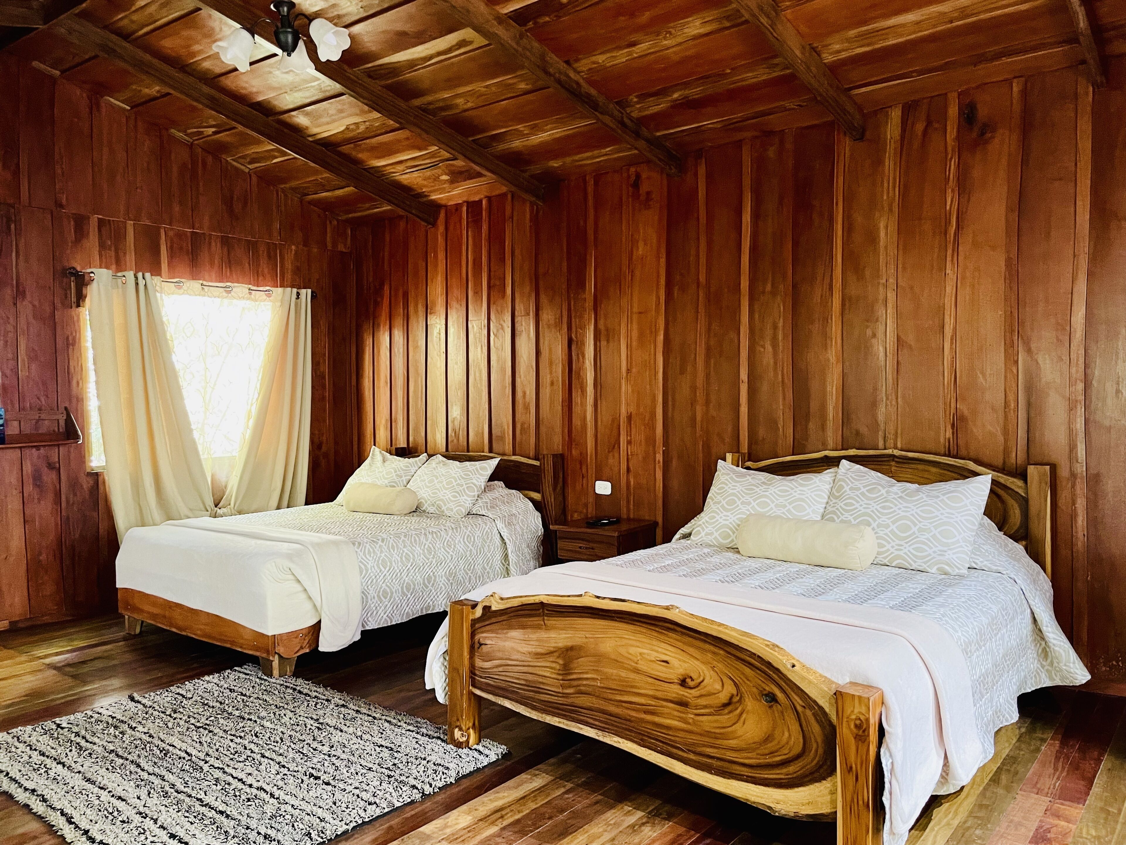 A wooden room with two beds, a nightstand, a lamp, and a window with curtains.