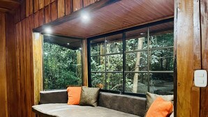 A wooden bench with cushions, a window with a view of greenery, and a light switch on the wall. - Cabañas La Montaña (Monteverde)