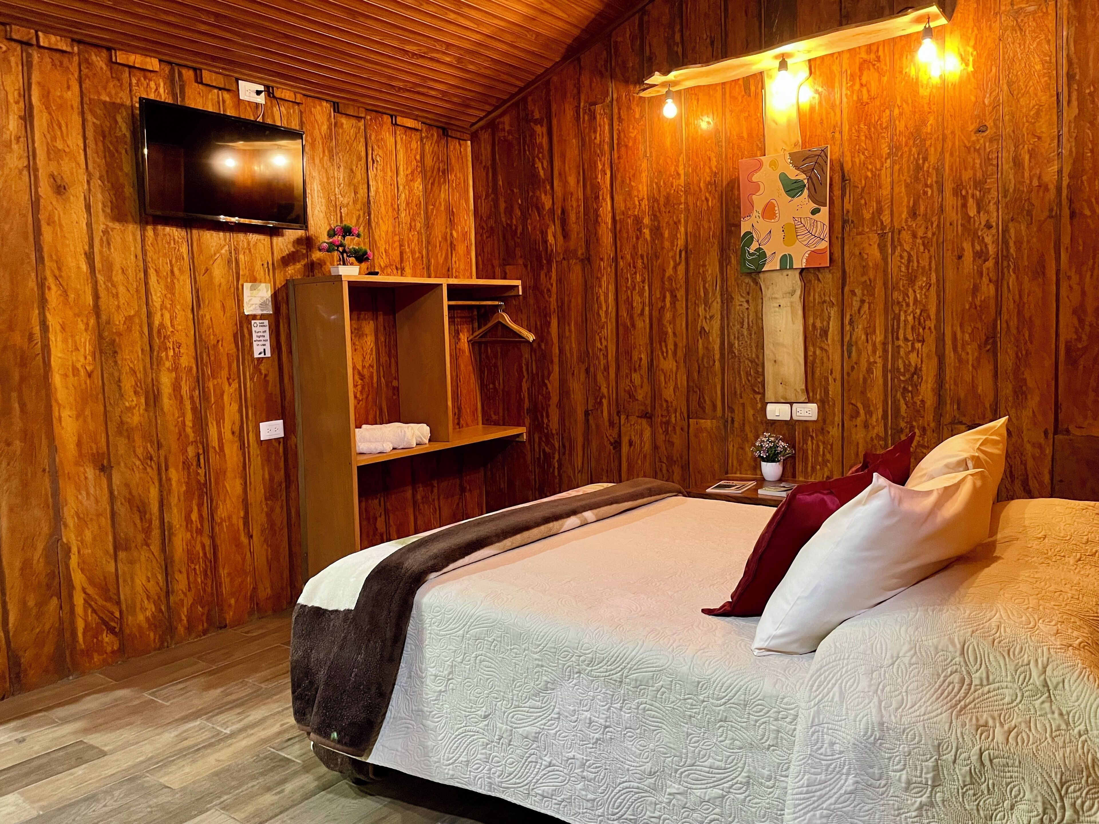 A wooden-paneled room with a bed, a TV mounted on the wall, and a shelf with towels.
