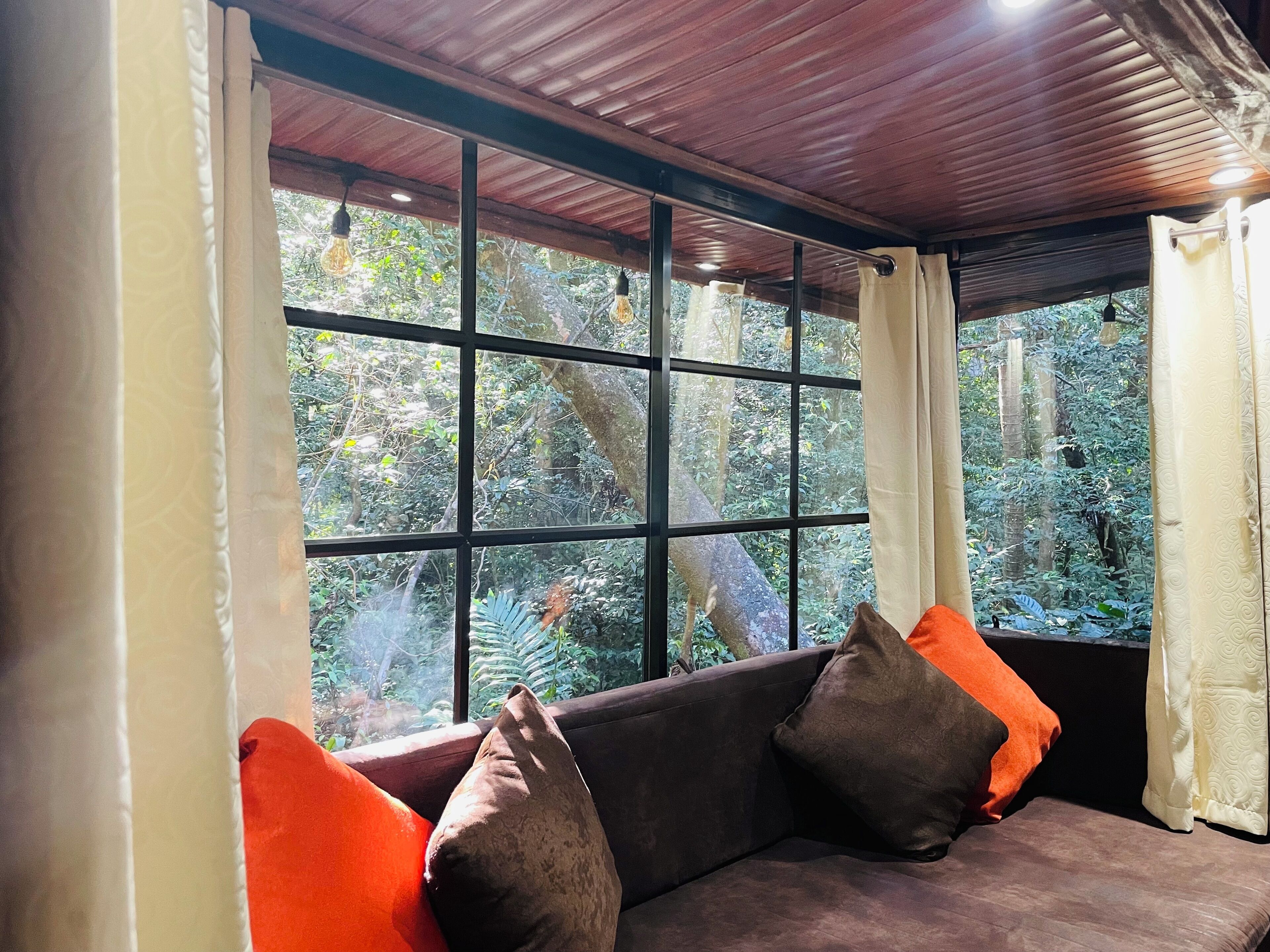 A room with a wooden ceiling, a cushioned sofa with orange and brown cushions, and a large window with curtains.