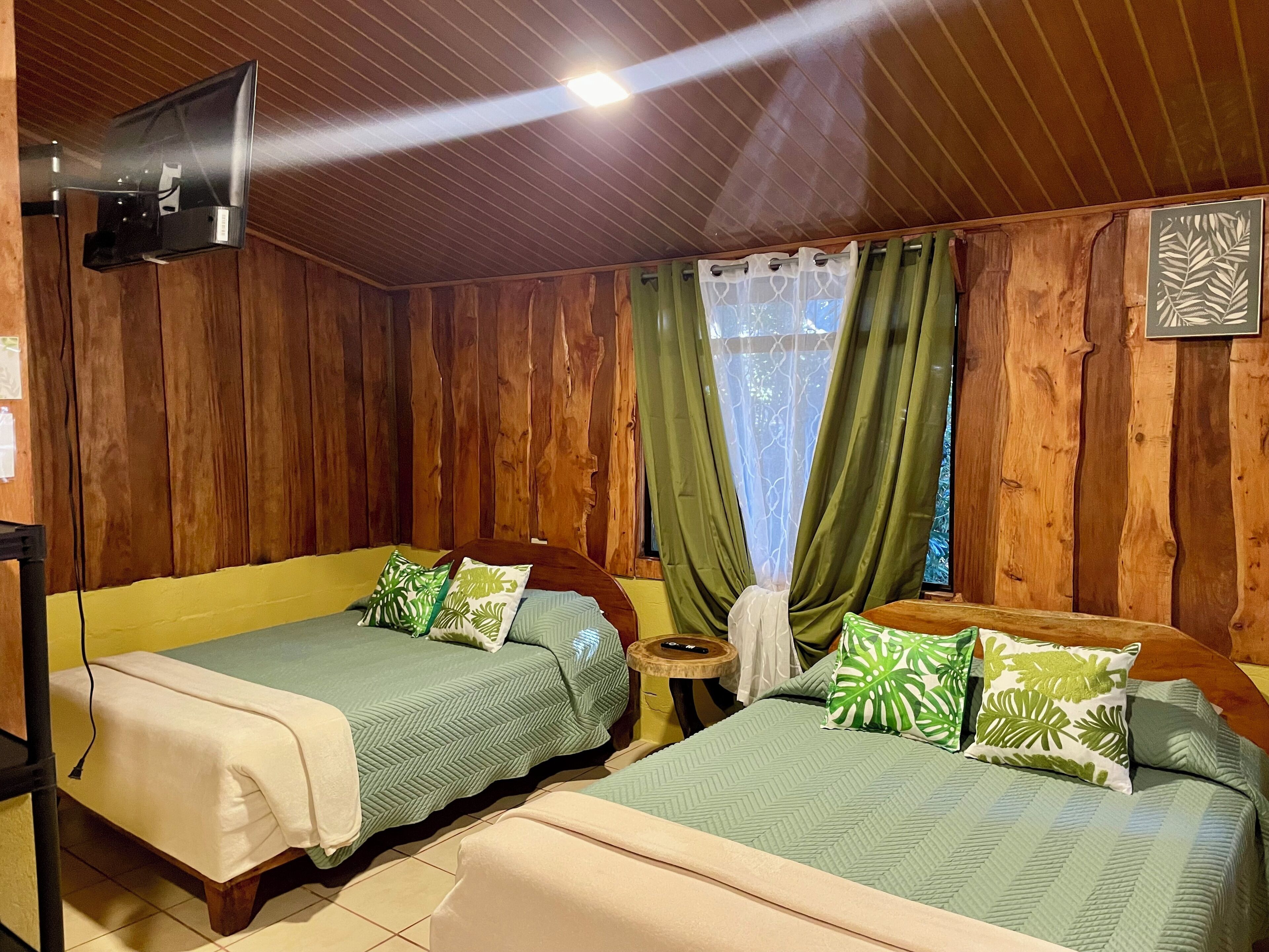 A room with two beds, wooden walls, a small table, a TV mounted on the wall, and a window with curtains.