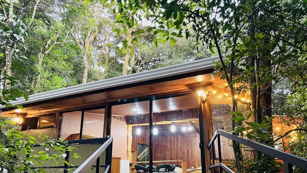 A modern, rustic cabin with a wooden exterior, large windows, and a cozy interior featuring a bed, seating area, and a dining space. - Cabañas La Montaña (Monteverde)