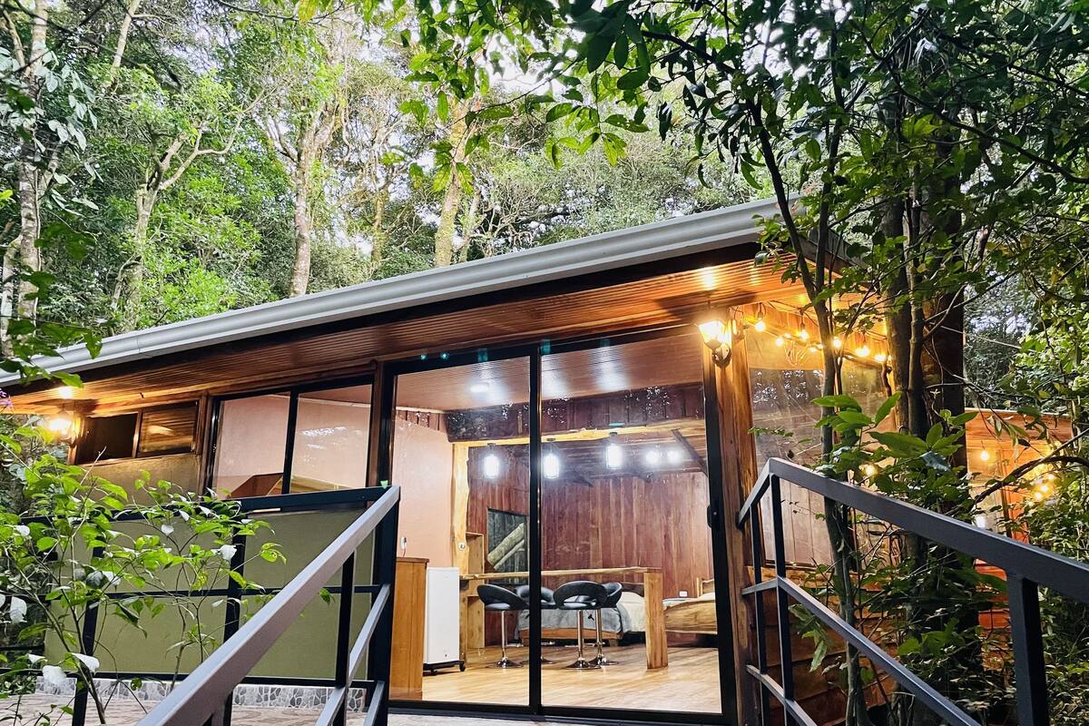 A modern, rustic cabin with a wooden exterior, large windows, and a cozy interior featuring a bed, seating area, and a dining space.