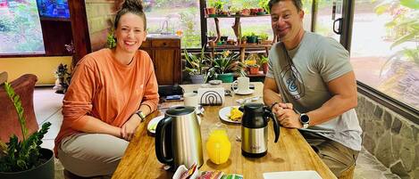 Un comedor con mesa de madera, dos personas sentadas y varios objetos como una tetera, una cafetera y comida.