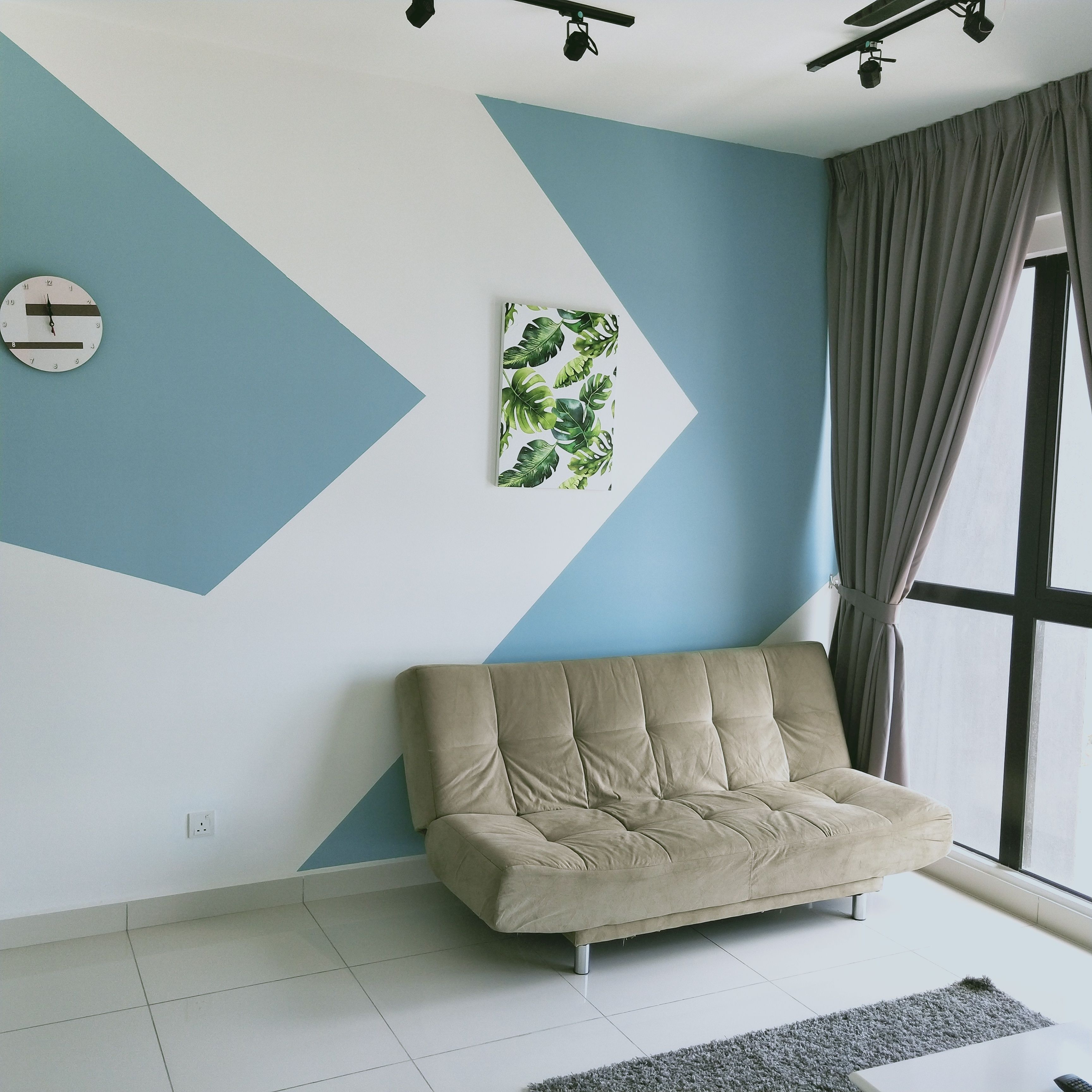 A modern living room with a beige sofa, a geometric wall design, a clock, and a window with curtains.