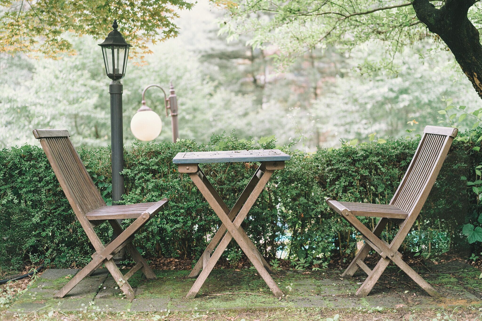 Dos sillas de madera y una mesa en un jardín, con una farola y follaje.