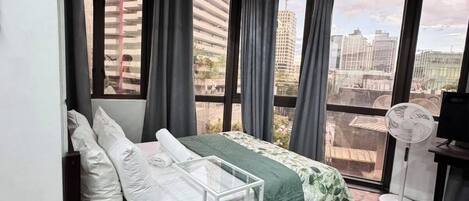 A room with a bed, a standing fan, and large windows with curtains.