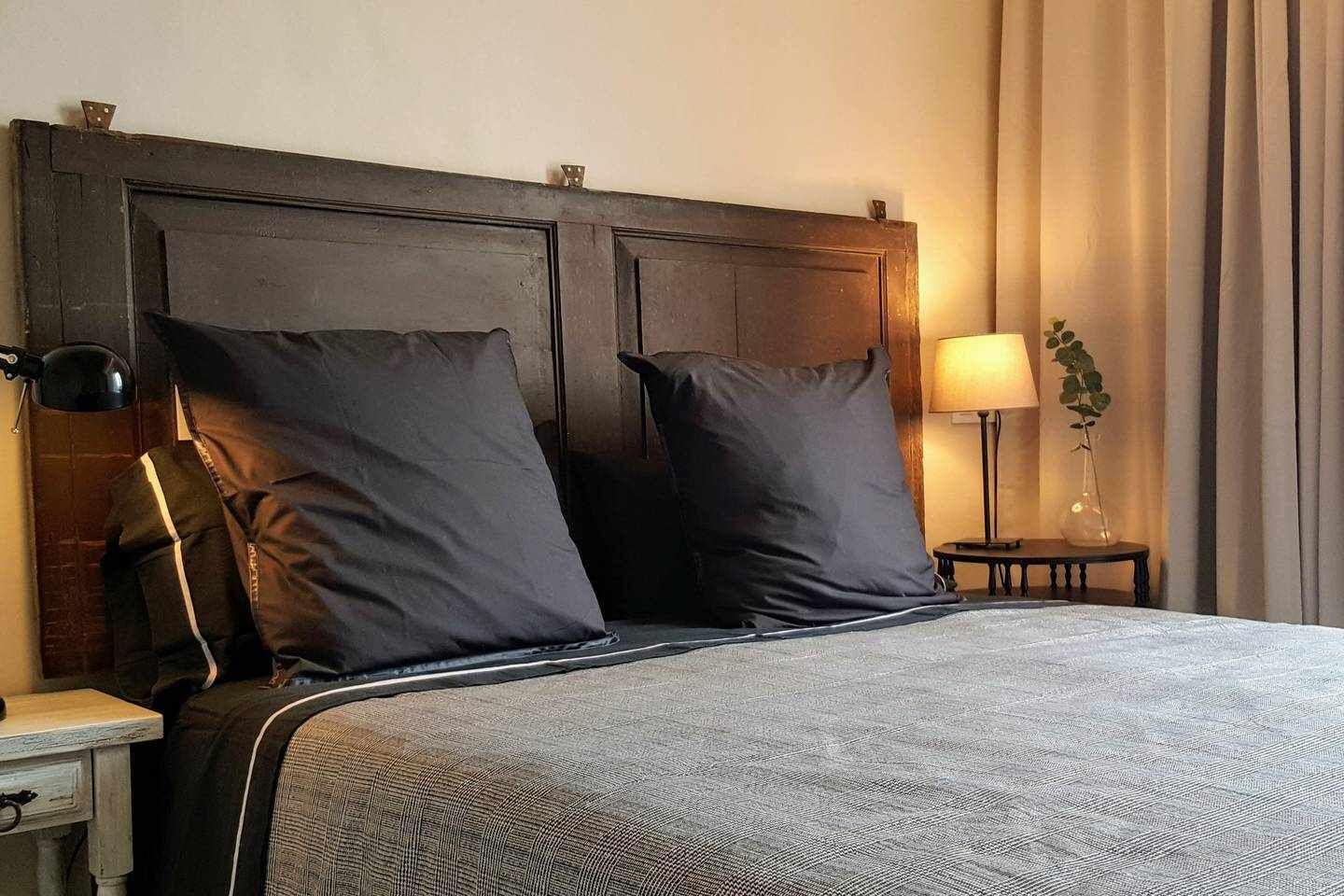 Un lit bien fait, avec une literie aux couleurs sombres, une lampe de chevet et une petite table sur laquelle est posée une plante.