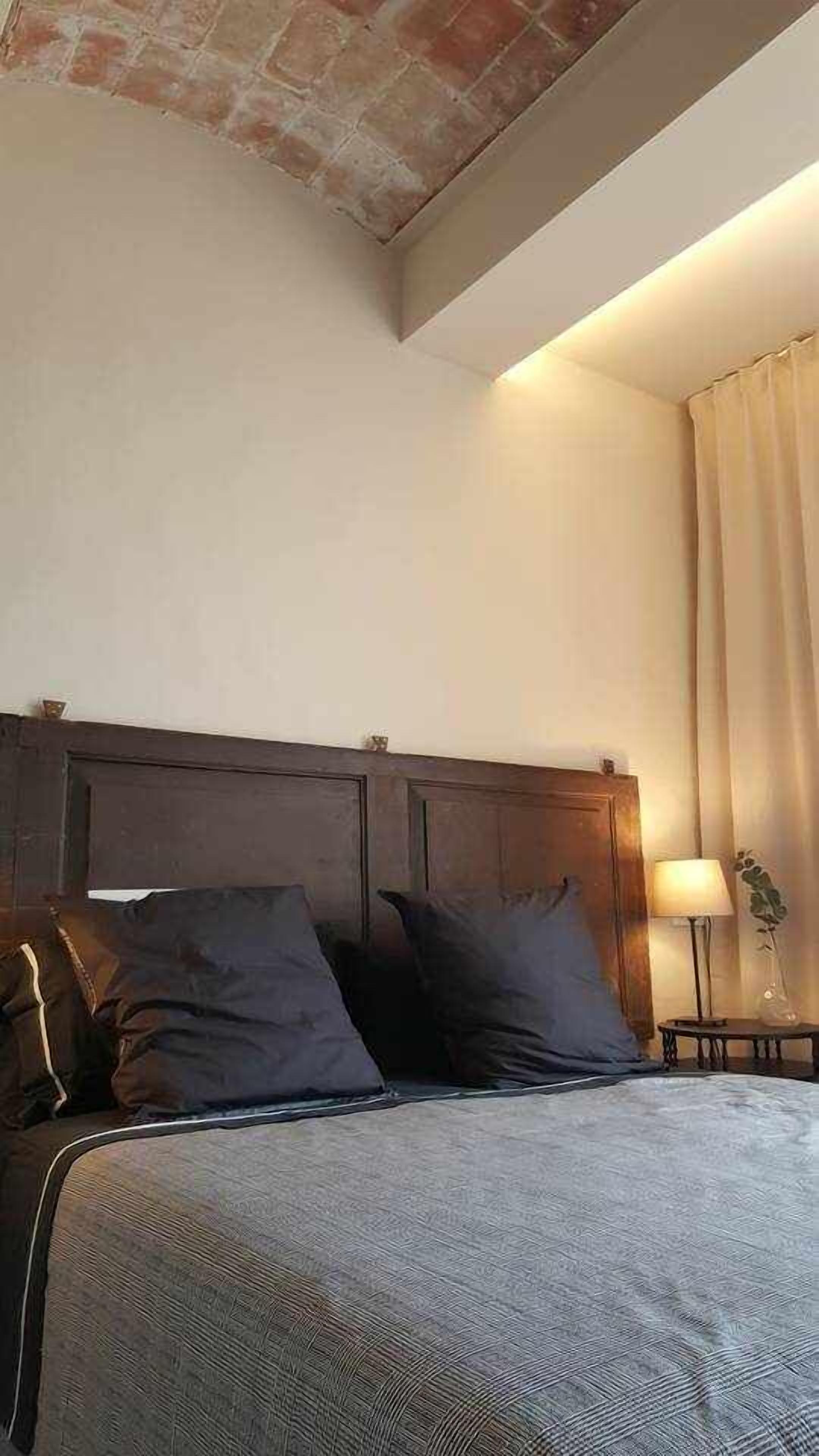 Un dormitorio con cabecera de madera, cama con ropa de cama gris, mesita de noche con lámpara y ventana con cortinas.