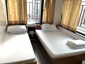 A room with two single beds, each with white linens and pillows, a small table with a stack of towels, and a window with curtains.