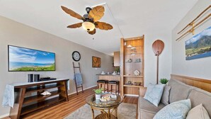 Una sala de estar moderna con televisor, muebles de madera, ventilador de techo y una zona de cocina.