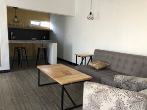 Ein modernes Wohnzimmer mit grauem Sofa, einem Holztisch und einem gemusterten Sessel. Im Hintergrund ist eine Küchenzeile mit einem Barhocker und Pendelleuchten zu sehen.