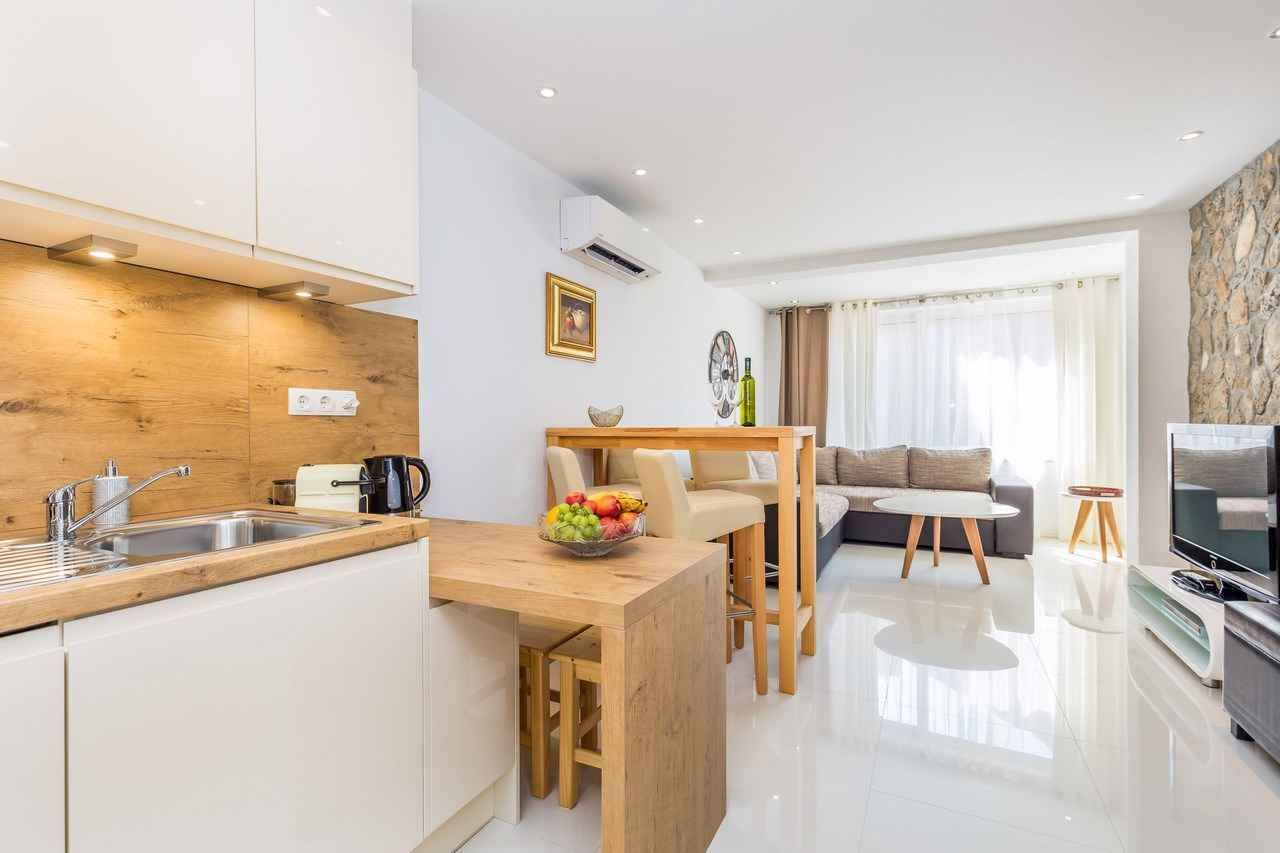 Una cocina moderna con encimera de madera, zona de comedor con mesa y sillas, sala de estar con sofá y televisión, y una pared de acento de piedra.